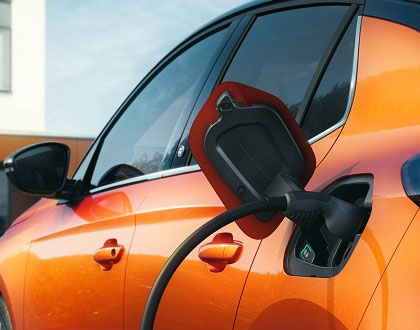 Ein oranges Elektroauto wird aufgeladen. Ein Ladekabel ist an den Ladeanschluss an der Seite des Fahrzeugs angeschlossen. Das Auto steht in der Nähe eines weißen Gebäudes.