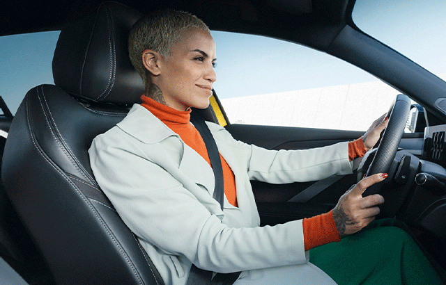 Eine Person mit kurzem, blondem Haar sitzt lächelnd am Steuer eines Autos. Sie trägt einen weißen Mantel über einem orangefarbenen Rollkragenpullover und hat ihre Hände am Lenkrad. Die Umgebung ist minimalistisch, mit einem hellen Himmel im Hintergrund.