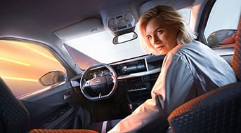 Eine Person mit kurzen blonden Haaren sitzt auf dem Fahrersitz eines Autos und blickt zurück über die Schulter. Das Innere des Autos zeigt ein modernes Armaturenbrett und ein Lenkrad. Durch die Fenster ist ein heller, verschwommener Hintergrund zu sehen.